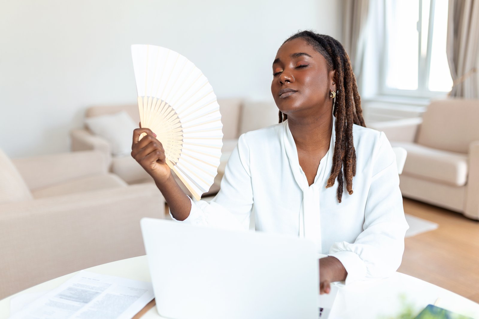 tired african woman work on laptop at home with a fan too hot from hot flashes in perimenopause