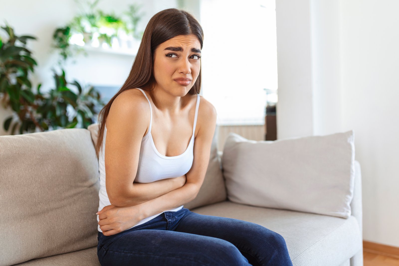 woman in painful expression holding hands against belly suffering menstrual period pain, lying sad on home bed, having tummy cramp in female health concept
